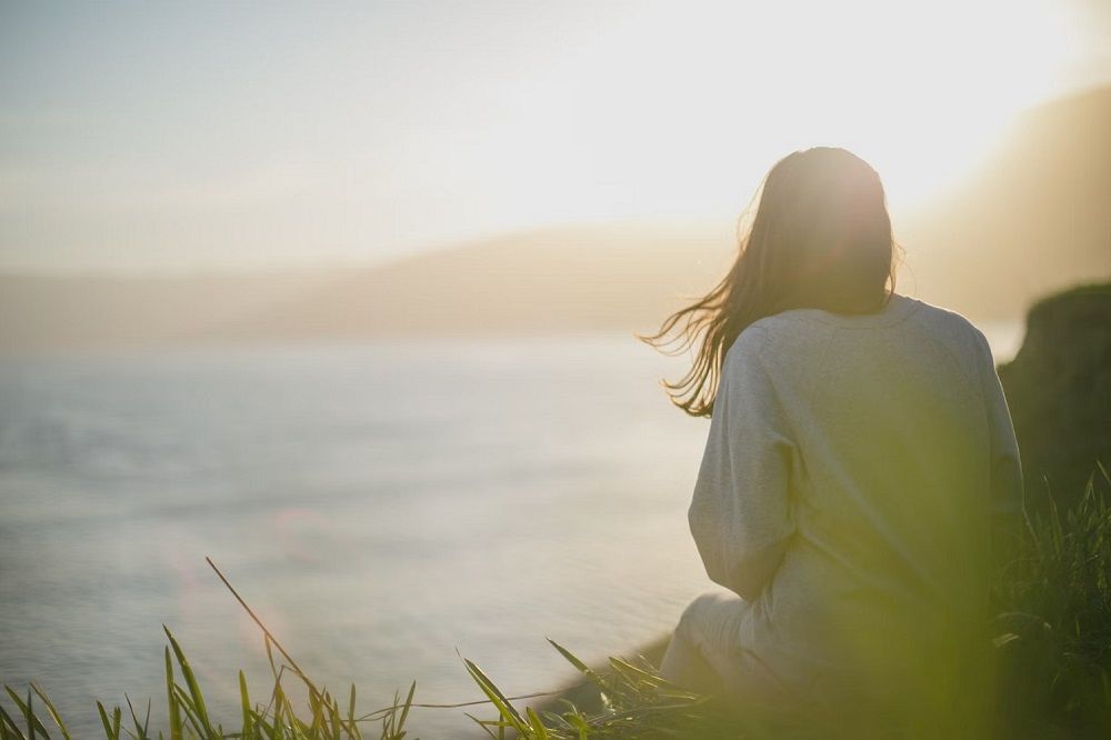 A woman perched on a rock, contemplating the serene ocean view, as the sun reflects off the water's surface.